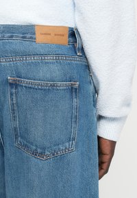 Jean en denim bleu avec une étiquette en cuir SAMSØE SAMSØE, présentant une poche arrière et des surpiqûres contrastées. Texture douce et coupe décontractée.