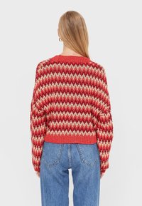 Pull tricoté rouge et beige avec un motif en zigzag, design cropped et encolure côtelée. Porte avec un jean bleu taille haute.