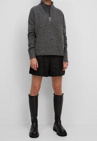 Persona che indossa un maglione grigio con zip, pantaloni corti neri e stivali in pelle neri fino al ginocchio su uno sfondo bianco semplice.