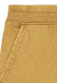 Gros plan sur la taille élastique et la couture de la poche latérale d'un pantalon ou short en tissu jaune moutarde.
