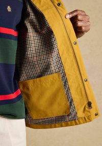 Hand die een opengele jas vasthoudt met een geruite binnenvoering en een grote binnenzak, gedragen over een meerkleurig gestreept shirt en witte broek.