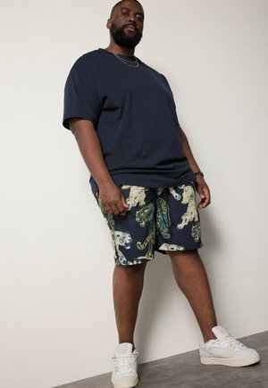 Mand iført en mørk oversized t-shirt, mønstrede shorts med tigre, hvide sneakers og en halskæde, står mod en ensfarvet lys væg.