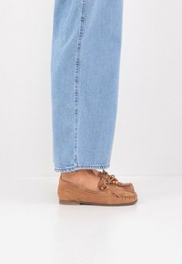 Chaussures mocassins en daim marron avec détail en franges, lacets et accents de perles en bois, associées à un jean en denim bleu clair.