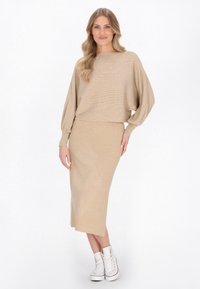 Beige gebreide trui met ballonmouwen en een cropped ontwerp, gecombineerd met een bijpassende rok tot de knie en witte hoge sneakers.