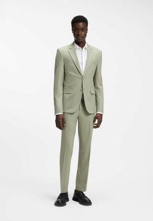 Persona alta vistiendo un traje de color verde salvia claro con una camisa blanca y zapatos negros, de pie contra un fondo blanco liso.