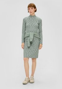 Sukienka dzianinowa w stonowanym zielonym kolorze z wysokim dekoltem i długimi rękawami, z paskiem związanym w talii, zestawiona z beżowymi sneakersami.
