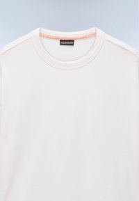 Hvid T-shirt med rund hals, orange syning på indersiden af kraven og sort mærke på lyseblå baggrund.