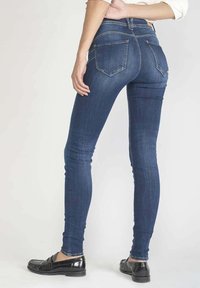 Jean skinny en denim bleu foncé avec une taille haute, comprenant deux poches arrière et des détails de couture subtils. Associé à des mocassins noirs.
