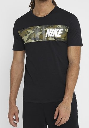 Mann trägt ein schwarzes Nike-T-Shirt mit einem horizontalen grünen und braunen Tarnstreifen über der Brust mit weißem Nike-Logo.