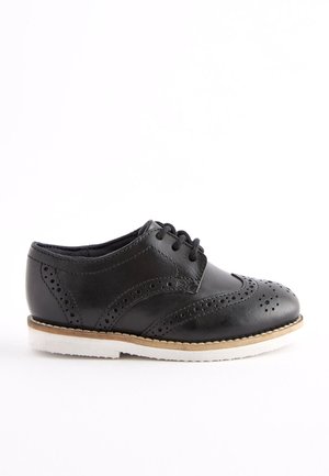 Sort læderbroguesko med dekorative perforeringer, sorte snørebånd og hvid sål, vist i profil på en enkel hvid baggrund.