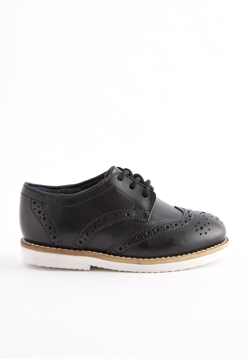 Chaussure brogue en cuir noir avec perforations décoratives, lacets noirs et semelle blanche, présentée de profil sur fond blanc uni.
