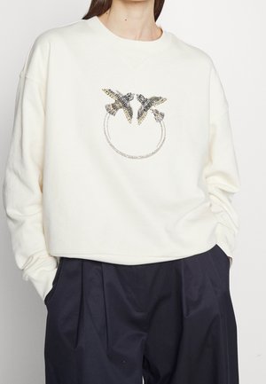 Femme portant un sweat-shirt blanc avec deux oiseaux ornés de bijoux se faisant face et un motif circulaire en chaîne en dessous, associé à un pantalon noir.