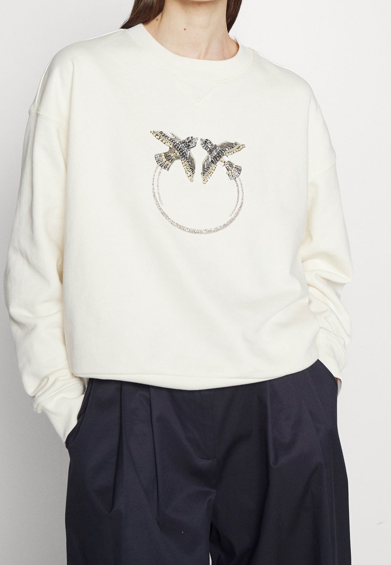 Femme portant un sweat-shirt blanc avec deux oiseaux ornés de bijoux se faisant face et un motif circulaire en chaîne en dessous, associé à un pantalon noir.