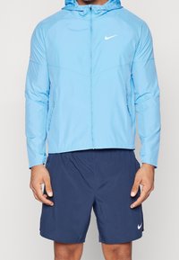 Veste zippée bleu clair avec capuche, en matière lisse, arborant un petit logo Nike blanc sur la poitrine. Portée avec un short marine.