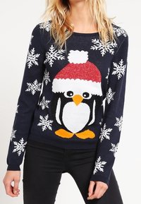 Modellen bär en marinblå tröja med en stor pingvin som har en röd hatt och snöflingemönster, tillsammans med svarta jeans.