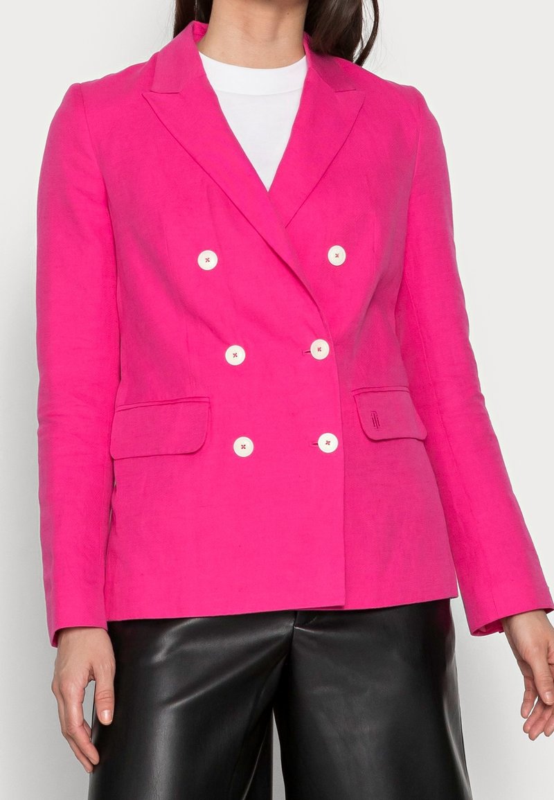 Femme portant un blazer rose vif à double boutonnage avec des boutons blancs sur un haut blanc et un pantalon en cuir noir.