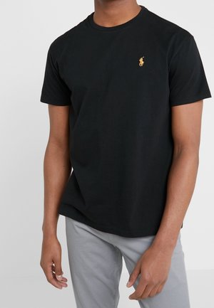 Een persoon draagt een zwart t-shirt met een klein gouden logo, staand tegen een effen witte achtergrond, met de handen naast zich.