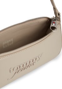 Μπεζ θήκη Tommy Jeans με ανάγλυφο λογότυπο στην πρόσοψη, ανοιχτό φερμουάρ που αποκαλύπτει λεπτομέρειες λογότυπου στο εσωτερικό και ασημί τράβηγμα φερμουάρ με χρώματα της μάρκας.