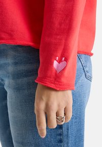 Hand entspannt an der Seite, trägt einen silbernen herzförmigen Ring, ein rotes Langarmoberteil mit kleinen rosa Herzstickereien an den Ärmeln und eine blaue Jeans.