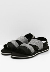 Mustat ja hopeiset kudotut tekstiilisaappaat, joissa on kaksi leveää säädettävää hihnaa. Ominaisuuksiin kuuluu tasapohja, jossa on hienovarainen valkoinen reuna.