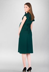 Zielona, długości do kolan sukienka ciążowa z krótkimi, falbanistymi rękawami i związanym w pasie obi belt. Gładki materiał z luźnym krojem, zestawiona z czarnymi szpilkami.