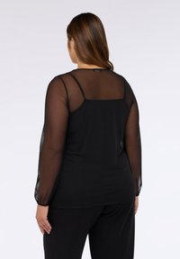Top nero con maniche lunghe trasparenti e scollatura arrotondata, realizzato in un materiale morbido con una texture liscia, abbinato a pantaloni neri.