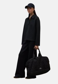 Mujer con una camiseta de manga larga negra, pantalones de pierna ancha y gorra, sosteniendo un gran bolso de viaje acolchado negro contra un fondo liso.