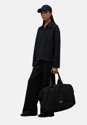 Mujer con una camiseta de manga larga negra, pantalones de pierna ancha y gorra, sosteniendo un gran bolso de viaje acolchado negro contra un fondo liso.