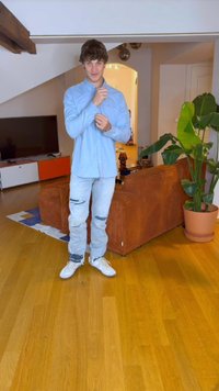 Svetlomodrá košeľa na gombíky, poškodené svetlé džínsy a biele tenisky, nosené v izbe s drevenou podlahou, pohovkou a veľkou rastlinou.