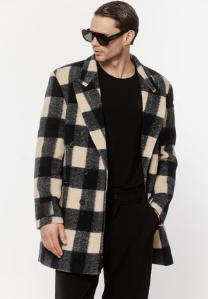 Mężczyzna w oversize'owym czarno-beżowym płaszczu w kratę, czarnej koszuli, czarnych spodniach i dużych czarnych okularach przeciwsłonecznych, patrzący na bok.