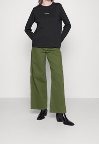 Felpa nera con logo Calvin Klein, abbinata a pantaloni verdi a gamba larga e stivali neri con tacco. Tessuto liscio e design aderente.