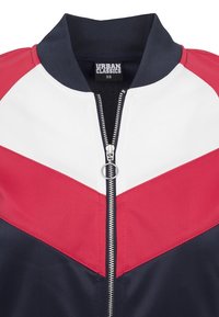 Rode, witte en marineblauwe windbreaker jas met een rits aan de voorkant, waarbij een chevronontwerp en een geribde kraag te zien zijn. Gemaakt van lichtgewicht stof.