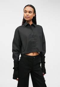 Cămașă neagră scurtă cu nasturi, cu mâneci lungi și guler, asociată cu pantaloni negri care au închidere cu fermoar și accente ribbate la încheieturi.