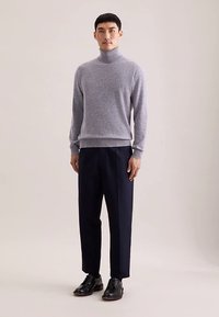 Hellgrauer Rollkragenpullover aus weichem Strickstoff, kombiniert mit dunkelblauen, maßgeschneiderten Hosen und schwarzen Anzugschuhen.