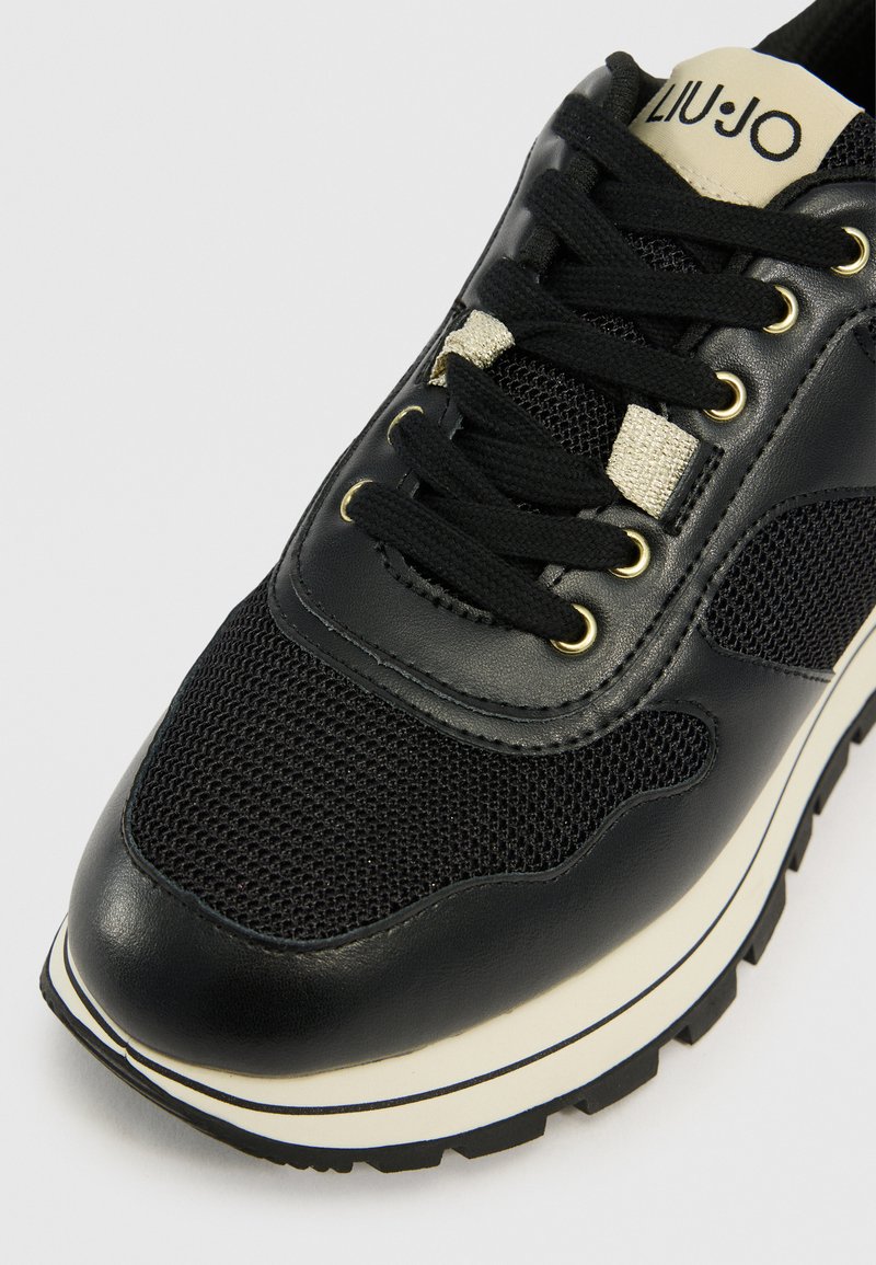 Schwarze Sneaker mit Obermaterial aus Leder und Mesh, goldakzentuierte Ösen, schwarze Schnürsenkel und dicke weiße Zwischensohle mit schwarzem Außensohlen. Markenname auf der Zunge.