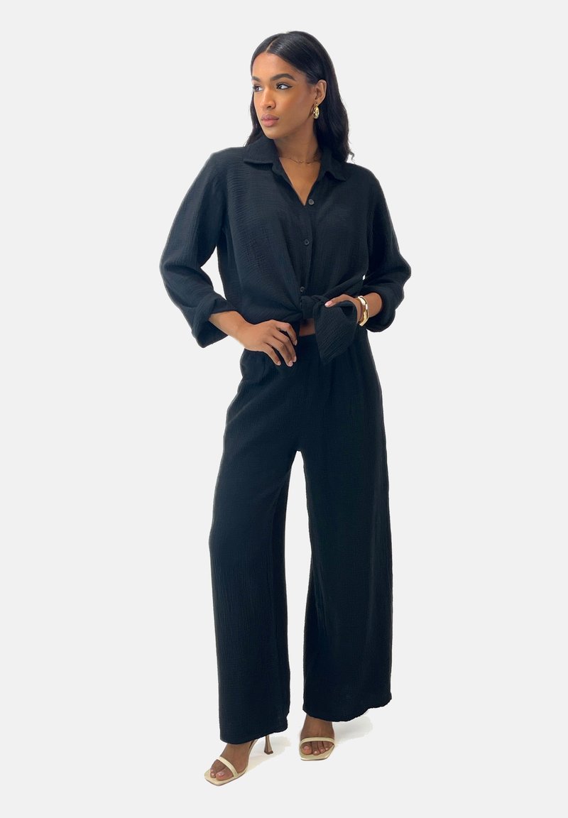 Zwarte tweedelige outfit bestaande uit een geknoopte blouse met kraag en wijde broekspijpen, gemaakt van een textuurstof, gecombineerd met open teen hakken.