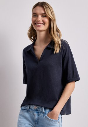 Lächelnde Frau mit blondem Haar trägt ein lockeres dunkelblaues kurzärmeliges Hemd mit Kragen und hellblaue Jeans, Hand in der Tasche, vor einfarbigem Hintergrund.