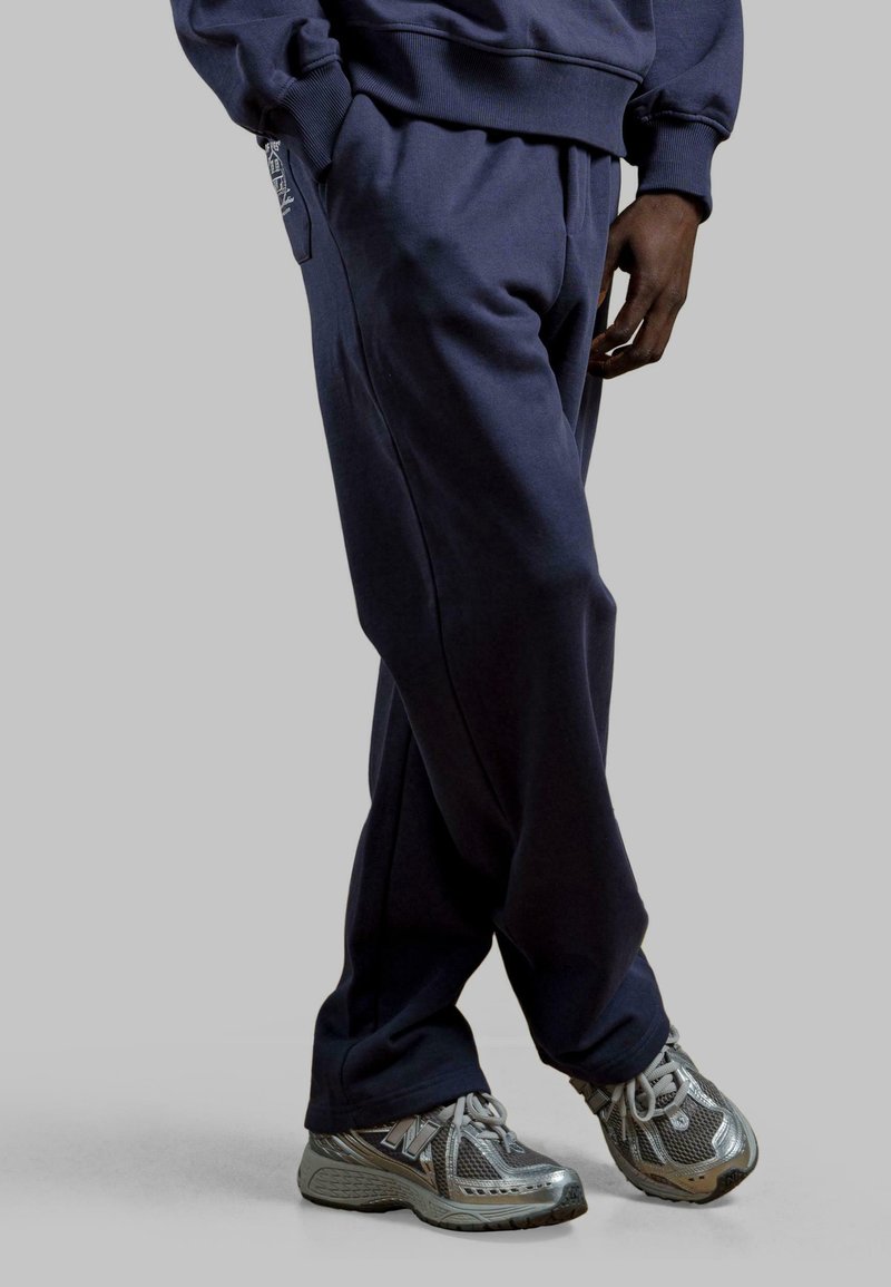Marineblauwe trainingsbroek van zacht materiaal met zijzakken, gecombineerd met grijze New Balance sneakers met een bovenkant van mesh en een gestructureerde zool.
