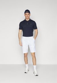 Granatowa koszulka polo z trzema guzikami i logo, zestawiona z białymi szortami. Model nosi białe sneakersy i granatową czapkę. Minimalistyczny design.