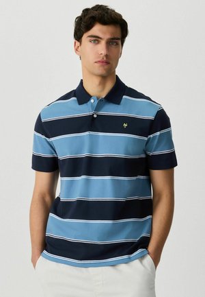 Junger Mann mit kurzen lockigen Haaren, der ein blaues und marineblau gestreiftes Poloshirt und weiße Hosen trägt, mit den Händen in den Taschen stehend.