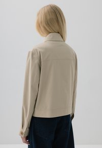 Beżowa krótka kurtka z wyprofilowanym kołnierzem, długimi rękawami i guzikami przy mankietach. Gładka tekstura, minimalistyczny design, zestawiona z ciemnoniebieskimi dżinsami.