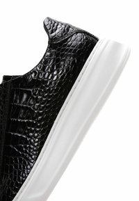 Sneaker in pelle nera con un motivo testurizzato simile a quello di un rettile, caratterizzato da una suola in gomma bianca e un design elegante. La vista laterale mette in evidenza la forma.