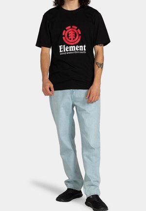 T-shirt en coton noir avec un logo Element rouge et blanc, associé à un jean denim bleu clair. Le modèle porte des chaussures de sport noires.