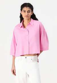 Korte ermer, cropped button-up skjorte i lys rosa stoff, med kragehals og avslappet passform. Bæres med hvite bukser.