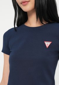 Tumedast sinise puuvillane T-särk ümmarguse kaelusega ja lühikeste varrukatega, mille rinnapiiril on väike kolmnurkne punane "GUESS" logo.