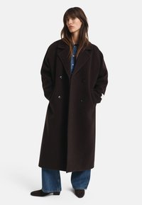Vrouw die staat met een lange donkerbruine dubbele coat over een paar blauwe jeans en donkere schoenen, met handen in de zakken van de coat tegen een witte achtergrond.