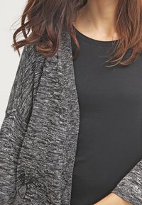 Mujer con cabello largo castaño que lleva una camisa negra y un cárdigan gris oscuro texturizado de frente abierto.