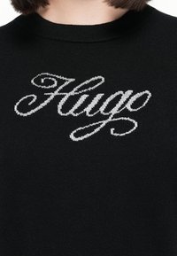Suéter negro de punto con un destacado logo plateado bordado "Hugo" en el pecho en una fuente cursiva.
