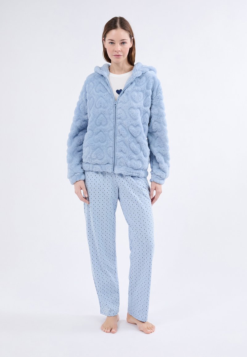 Mujer de pie descalza, vistiendo una sudadera de forro polar azul claro con un estampado de corazones y pantalones de pijama a rayas a juego, sobre un fondo blanco.