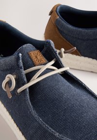 Blaue Leinenschuhe mit einem kontrastierenden braunen Wildlederabsatz. Sie haben hellgraue Schnürsenkel und einen strukturierten Lederfleck auf der Zunge. Weiße, gesprenkelte Sohle.